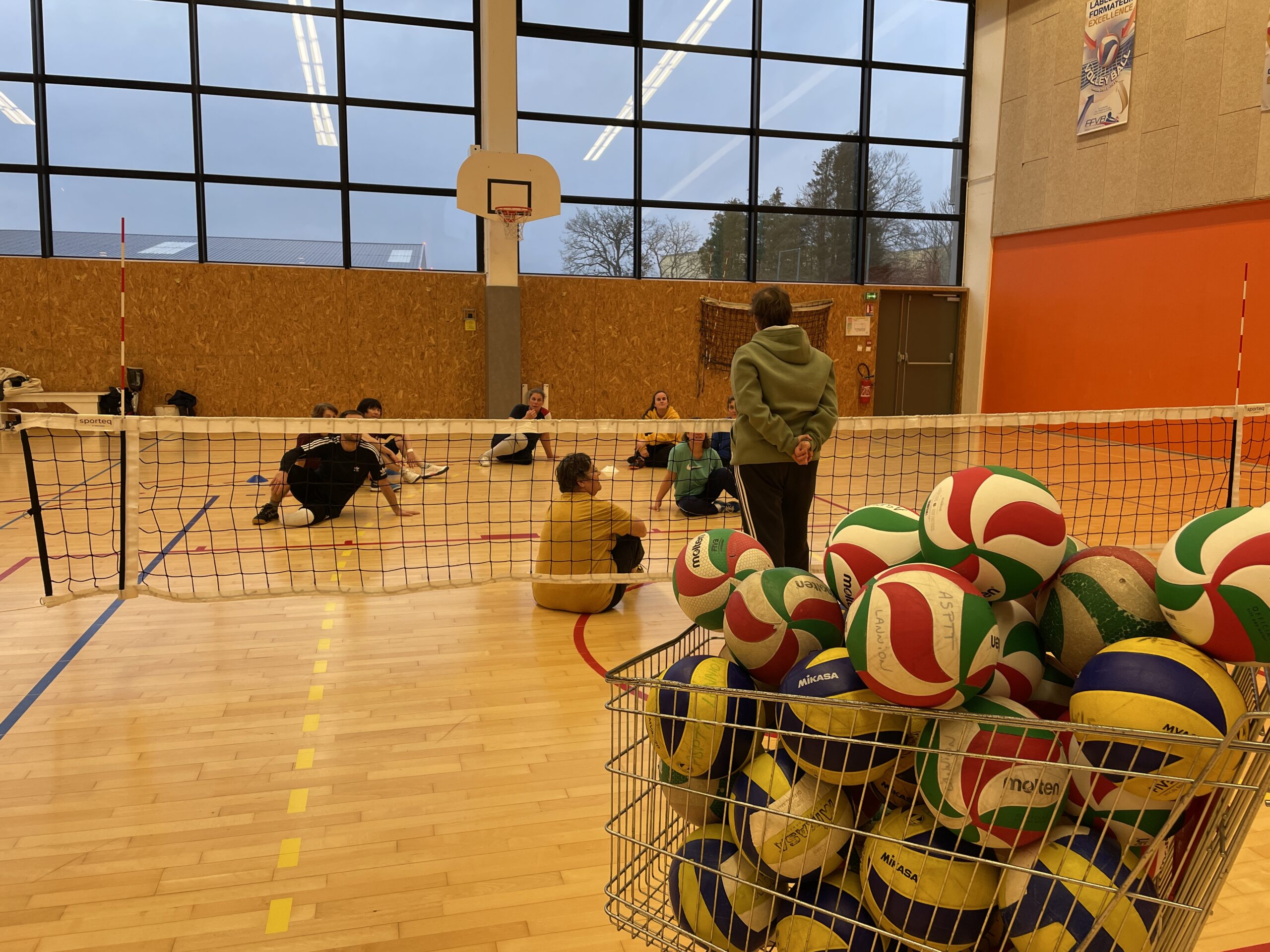 Les licenciés de l'ASPTT s'entrainent, Nathalie Prigent les observe, debout devant le filet. Au premier rang se trouve un caddie de ballons de volley.